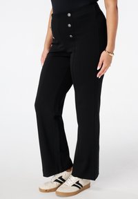 Vrouw in zwarte hooggehakte broek met zilveren knopen, witte sneakers met zwarte strepen en gouden armbanden om haar pols.