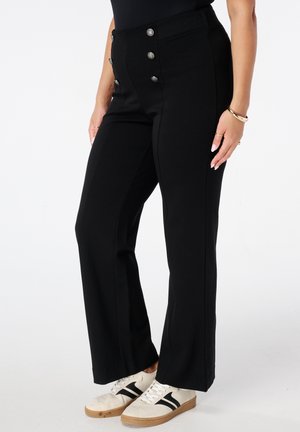 Vrouw in zwarte hooggehakte broek met zilveren knopen, witte sneakers met zwarte strepen en gouden armbanden om haar pols.