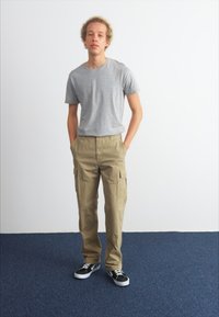 Grå kortärmad t-shirt, khakifärgade cargo-byxor med sidofickor och svarta sneakers på en modell mot en vit bakgrund och blå golv.