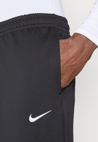 Μαύρο αθλητικό παντελόνι από λείο ύφασμα, με ελαστική μέση, πλευρική τσέπη και λευκό λογότυπο Nike.