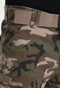 Die Tarn-Cargohose verfügt über eine Mischung aus grünen, braunen und schwarzen Mustern, mit einem robusten gewebten Gürtel und mehreren Taschen.