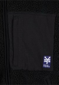 Svart fleecejakke med teksturert overflate, med en glatt stofflomme og en lilla Zoo York-logo-patch.