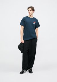 Σκούρο μπλε βαμβ�ακερό T-shirt με κοντά μανίκια, που διαθέτει κόκκινο λογότυπο στο αριστερό στήθος. Συνδυάζεται με φαρδιά μαύρα παντελόνια και μαύρα αθλητικά παπούτσια.