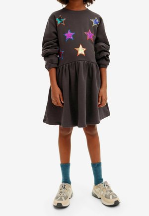 Niño con vestido negro de mangas remangadas y estrellas de lentejuelas multicolores, calcetines azules y zapatillas beige, de pie contra un fondo blanco.