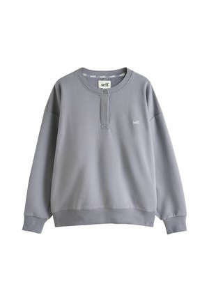 Blonde Frau mit welligem Haar, die einen grauen Sweatshirt und schwarze Shorts mit "self."-Logo trägt, steht mit Händen in Taillennähe im Sonnenlicht.