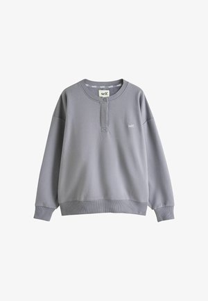 Lyshåret kvinde med bølget hår iført en grå sweatshirt og sorte shorts med "self." logo, stående med hænderne ved taljen i sollys.