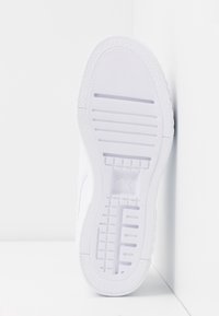 Semelle d'une basket blanche avec une adhérence texturée et des rainures, positionnée à la verticale contre un mur d'angle blanc sur une surface blanche.