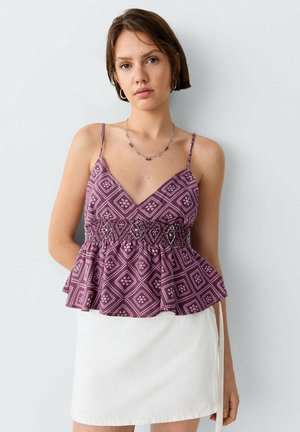 Femme portant un haut violet sans manches à motifs avec un ourlet volanté et une jupe blanche, debout devant un fond gris clair uni.