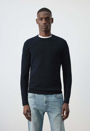Hombre con un jersey negro liso de manga larga sobre una camisa blanca y vaqueros azul claro, de pie frente a un fondo claro y liso.