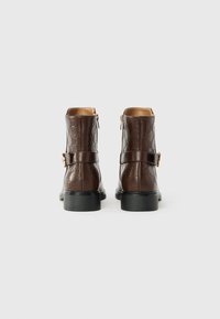 Bottines en cuir marron avec motif gaufré, fermeture éclair sur le côté, et sangle réglable avec boucle. Semelle noire plate. Texture lisse.