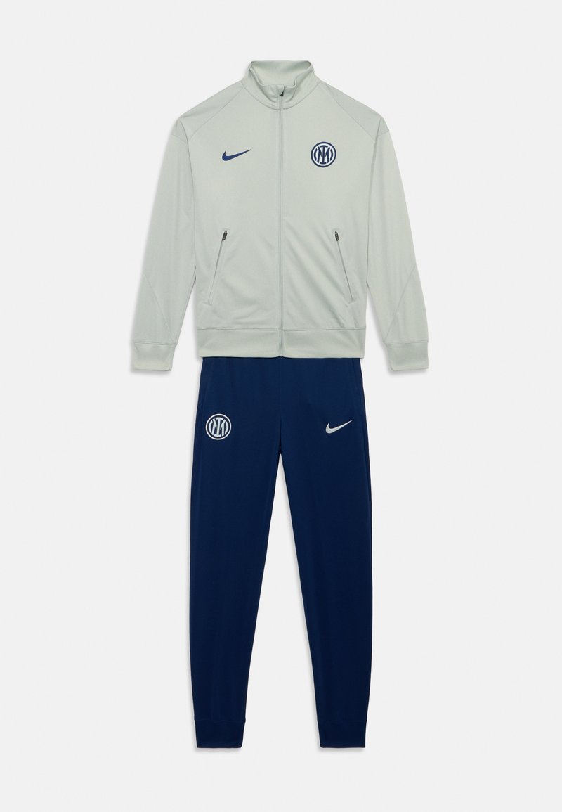 Veste zippée gris clair avec col montant, poches latérales, pantalon marine. Présente un logo bleu sur la poitrine et le pantalon. Tissu lisse, coupe athlétique.