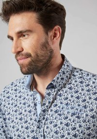 Chemise à motifs floraux bleus et blancs avec un col boutonné, présentant des accents contrastés bleu clair et un logo brodé sur la poitrine.