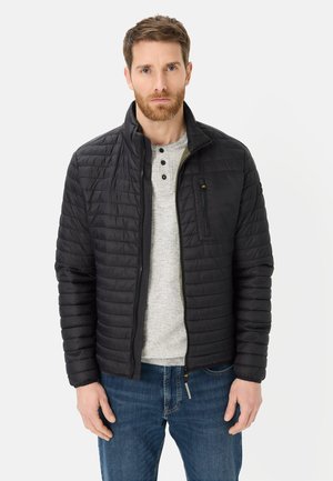 Mann trägt eine schwarze gesteppte Jacke über einem grauen Henley-Shirt und blauen Jeans, steht vor einem schlichten hellen Hintergrund.