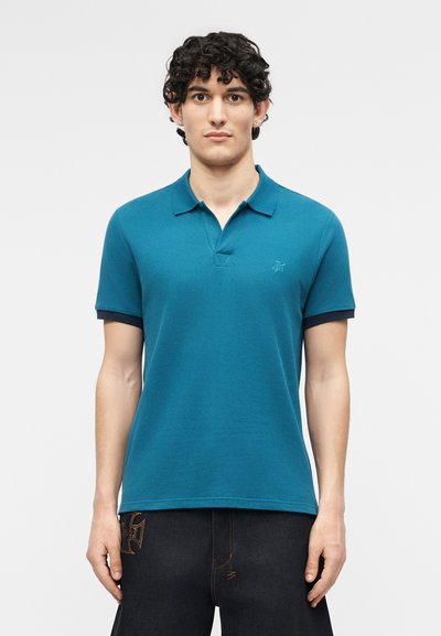 Homme aux cheveux bouclés portant un polo à manches courtes bleu sarcelle et un pantalon foncé devant un fond clair uni.