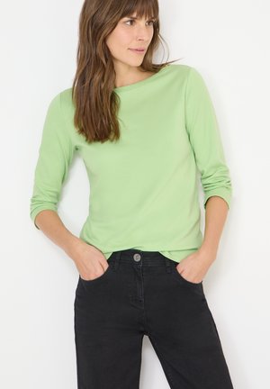 Haut vert clair à manches longues avec un col rond et des poignets retroussés, associé à un jean noir équipé d'un bouton argenté et de poches avant.