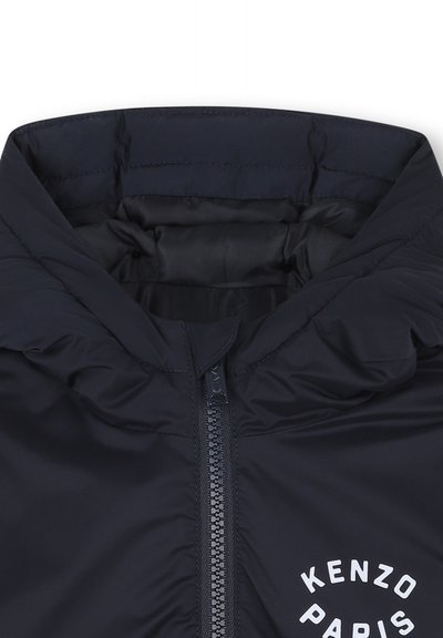 Giacca isolante blu navy con colletto alto, zip completa e logo circolare bianco con testo sul davanti. Texture del tessuto liscia e lucida.