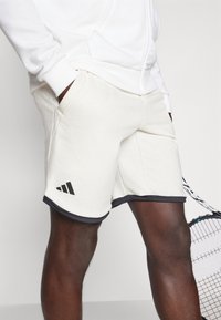 White athletic shorts with a black hem, made of soft fabric. Features front pockets and black three-stripe logo on the left leg.