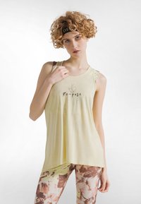 Lys gul tanktop med "Re-pose" trykt i sort. Har en rund halsudskæring og lagdelt skulderdesign. Kombineret med leggings med blomsterprint.