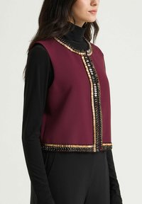 Veste sans manches bordeaux ornée d'embellissements métalliques le long de l'encolure et de l'ourlet, associée à un top noir ajusté à manches longues.