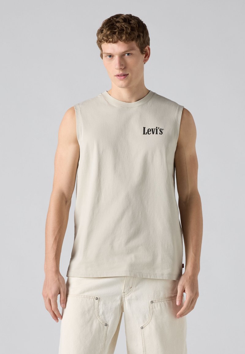 Jeune homme aux cheveux bouclés portant un débardeur beige Levi's et un pantalon clair assorti, debout devant un fond gris uni.
