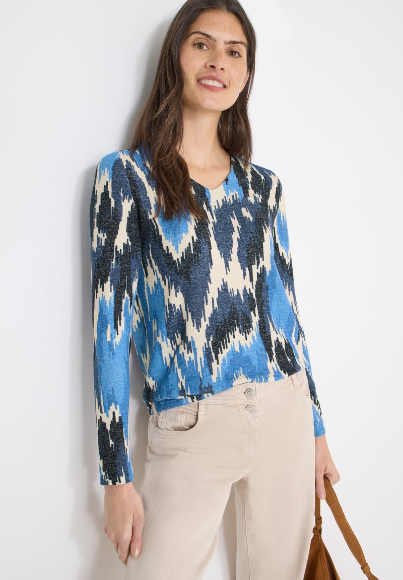 Pull à manches longues avec col en V, motif ikat en bleu, noir et crème ; tissu doux avec une coupe décontractée, associé à un pantalon beige clair et un sac marron.