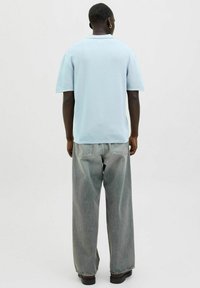 Lyseblå kortærmet polo-shirt med tekstureret finish, parret med løstsiddende grå jeans og sorte sko, set fra bagsiden.