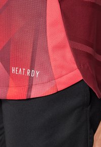 Rød idretts T-skjorte med teksturfinish, med gradientdesign og "HEAT.RDY" logo nær kanten. Kombinert med svarte shorts.