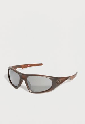 Rudos spalvos apjuostos saulės akiniai su tamsiais lęšiais ir Oakley logotipu ant šono, parodyti ant vientisos baltos fono.