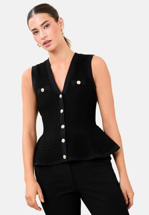 Vrouw draagt een zwarte mouwloze geribde top met zilveren knopen, zakken aan de voorkant en een peplumzoom, gecombineerd met een zwarte broek.