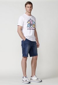 White cotton t-shirt with graphic mountain and bicycle design, paired with blue denim shorts, and white sneakers with gray accents.