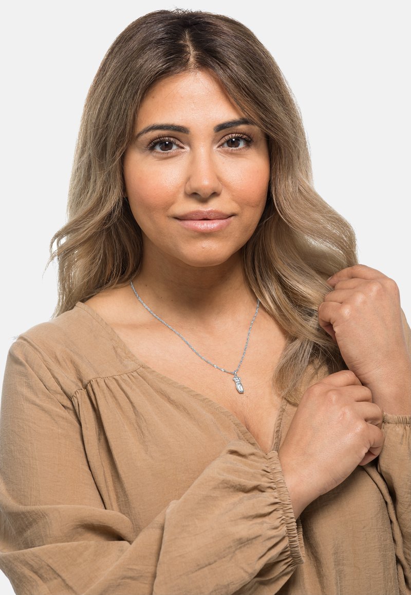 Srebrny naszyjnik z unikatowym wisiorkiem na cienkim łańcuszku, noszony na jasnobeżowej, teksturowanej bluzce z marszczonymi rękawami.