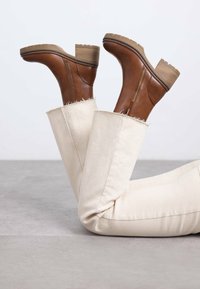 Botas de cuero marrón hasta el tobillo con suelas gruesas y tacones apilados, combinadas con jeans crema que tienen dobladillos deshilachados y una textura suave.
