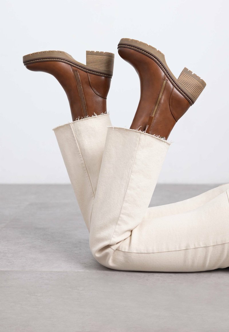 Botas de cuero marrón hasta el tobillo con suelas gruesas y tacones apilados, combinadas con jeans crema que tienen dobladillos deshilachados y una textura suave.