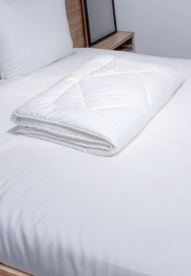 Weiß gesteppte Bettdecke, gefaltet auf einem weißen Bettüberwurf, mit glatter Textur und geometrischem Muster. Das Bett verfügt über ein Holz-Kopfteil und einen Nachttisch.