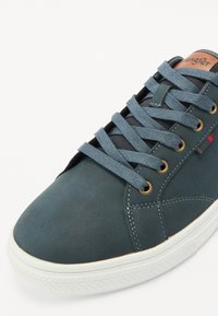 Sapatilhas de couro teal escuro com ilhós dourados, atacadores cinzas e sola de borracha branca. Apresenta detalhes cosidos e um logótipo na língua.