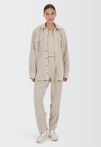 Beige corduroy shirt jacket with gold buttons, paired with matching ribbed top and pants. Model wears light sneakers, standing against a white background.