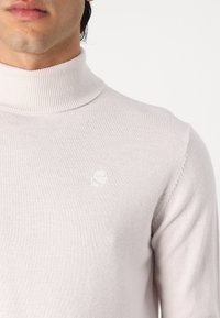 Hellgrauer Rollkragenpullover aus feinem Strickstoff. Verfügt über ein kleines weißes Logo auf der Brust und einen gerippten Halsausschnitt.