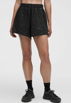 Vrouw die zwarte, getextureerde sportshorts draagt met een elastische tailleband, zwarte enkelsokken en zwarte hardloopschoenen tegen een effen achtergrond.