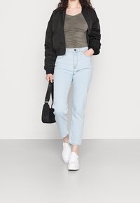 Schwarze Bomberjacke, olivfarbener, figurbetonter Oberteil, hellblaue, hochgeschnittene Jeans und weiße Sneaker. Schwarze Handtasche mit Umhängegurt. Lässiges Outfit.