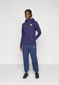 Lila Kapuzenjacke mit Reißverschluss, ausgestattet mit einem gelben Nike-Logo und dem FC Barcelona-Wappen. Marineblaue Jogginghose mit pinkem Nike-Logo und schmal zulaufenden Bündchen.