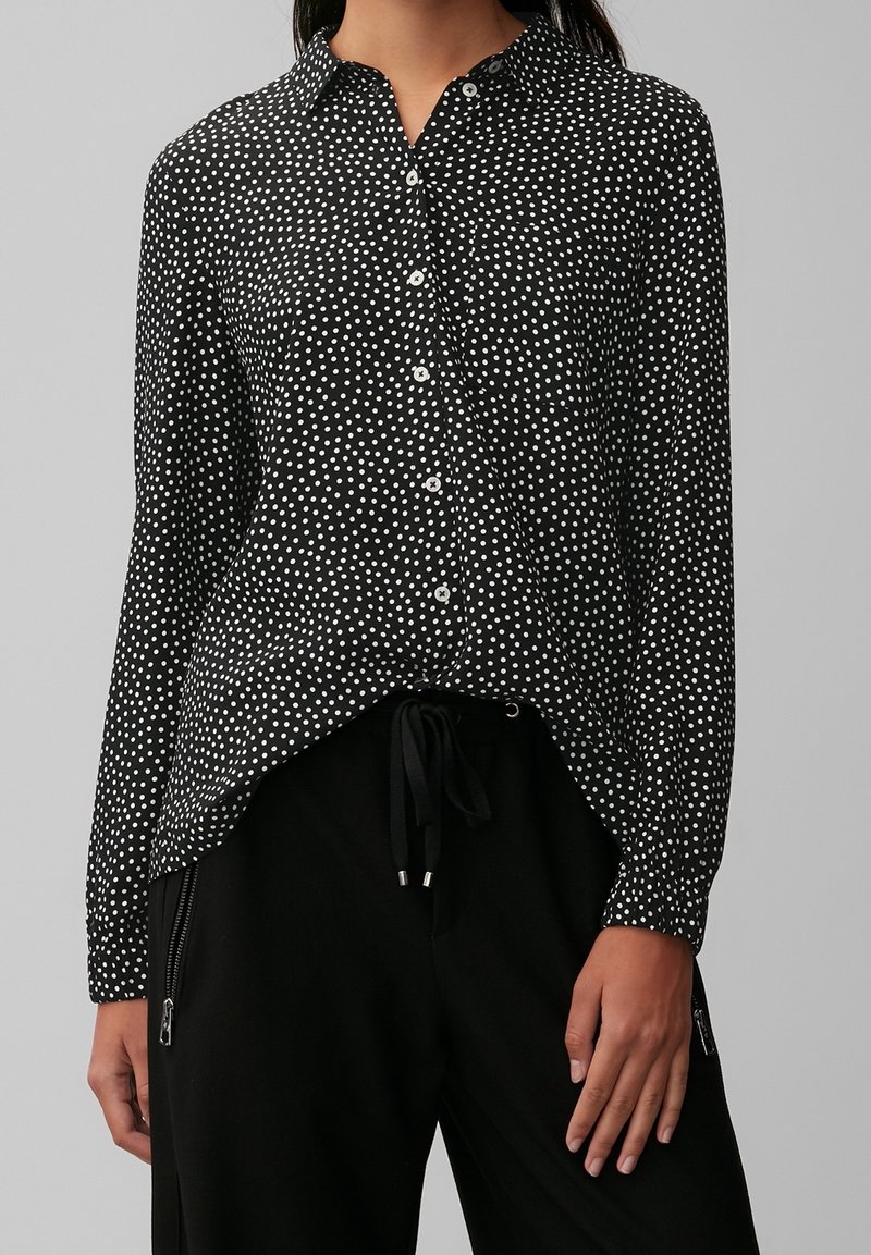 Femme portant une chemise noire à manches longues avec des pois blancs et un pantalon noir avec cordon de serrage et poches zippées.