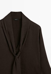 Blouse marron foncé à manches longues avec un col en V profond et un nœud de cravate attaché, en tissu texturé.