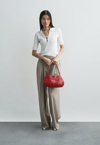 Geweven rode handtas met een gebogen vorm, korte handgreep en glanzende textuur. Model draagt een witte geribbelde top met kraag en beige wijde broek.