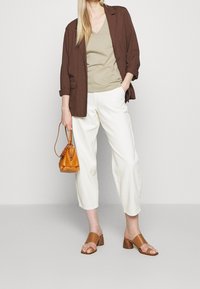 Blazer marrón sobre una blusa de cuello en V beige, pantalones blancos recortados y sandalias de deslizar marrones. Acompañado de un pequeño bolso naranja.