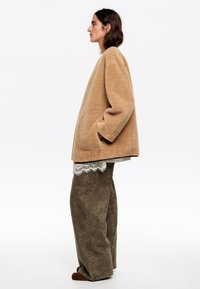 Chaqueta oversized beige y texturizada con bolsillos laterales. Llevada sobre una blusa con encaje y combinada con pantalones marrones sueltos y zapatos marrones.