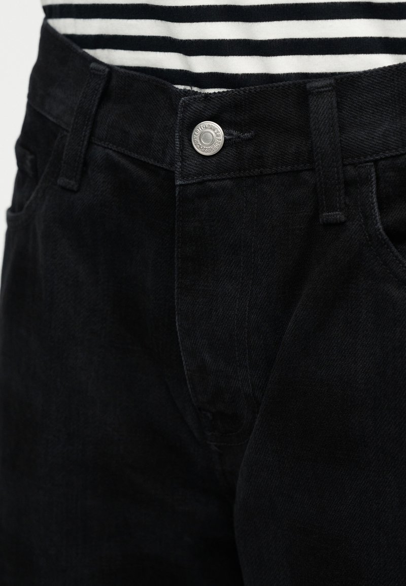 Jeans in denim nero con design aderente, chiusura con bottone argento, texture sottile e tasche laterali. Camicia a righe visibile sopra.