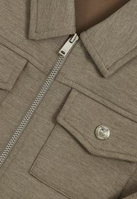 Taupe Zip-Jacke mit glatter Textur, ausgestattet mit einem metallischen Reißverschluss, aufgesetzter Tasche und Druckknopfdetails.