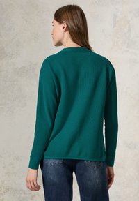 Teal ribbad tröja med långa ärmar och rundad halsringning, i kombination med mörka denimbyxor. Texturerat tyg med sidofickor i fållen.