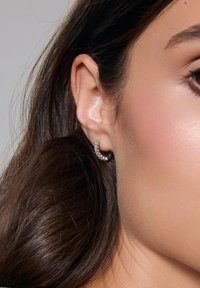 Srebrny kolczyk w kształcie obręczy z prostokątnymi i okrągłymi przejrzystymi kamieniami, charakteryzujący się polerowaną powierzchnią i zakrzywionym kształtem, zaprezentowany na rozmytym tle.