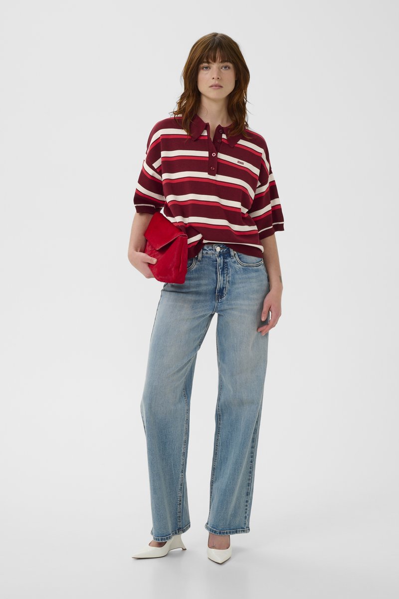 Polo rayé bordeaux et blanc avec col, associé à un jean bleu clair à jambes larges. Tient une pochette rouge et porte des escarpins blancs pointus.